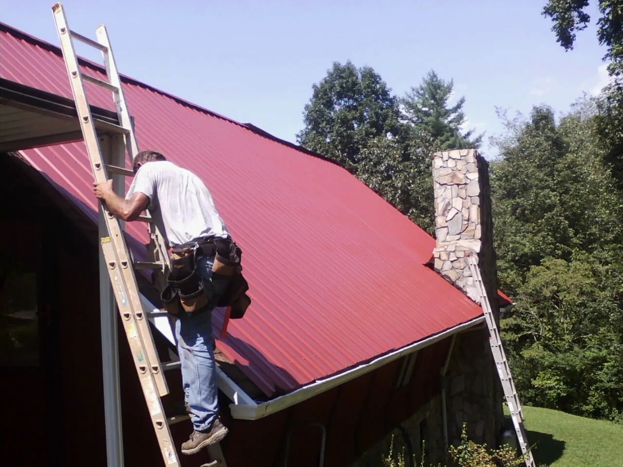 Roofer on a ladder beside a red metal roof and stone chimney — Soffit & Fascia Repair by Roofing Brothers in Cedar Hill